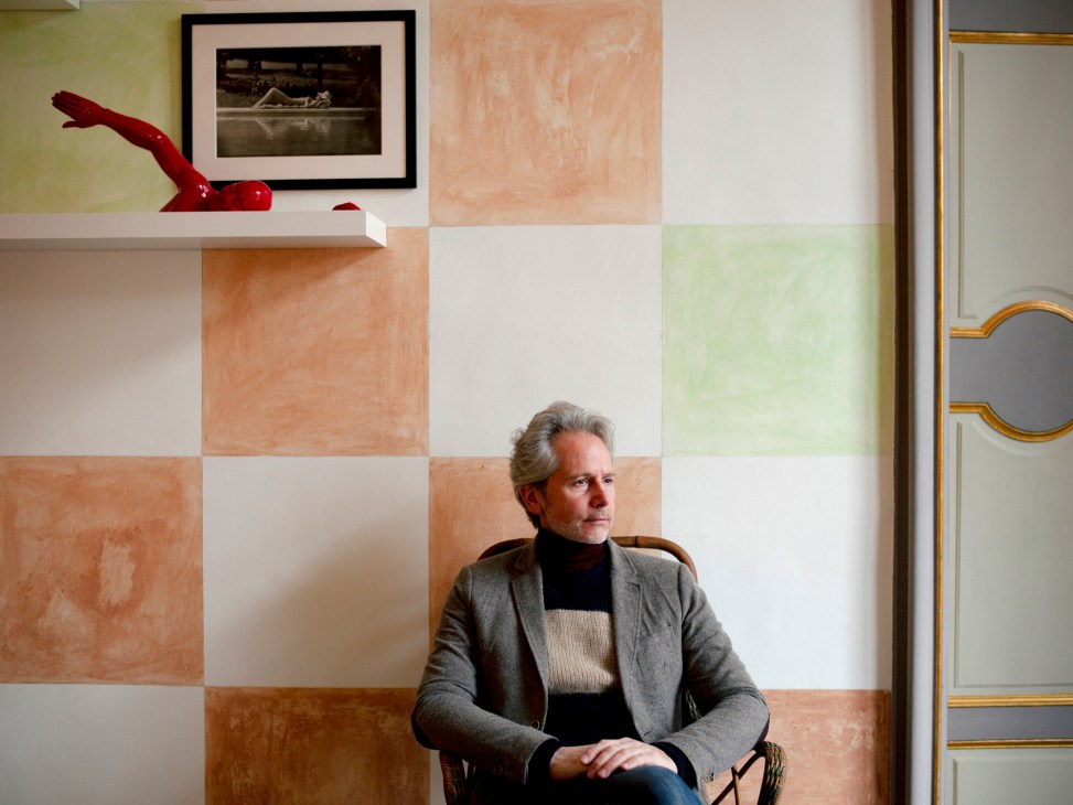 Middle-aged man with grey hair in grey blazer sits in chair against colorful checkered wall in elegant interior