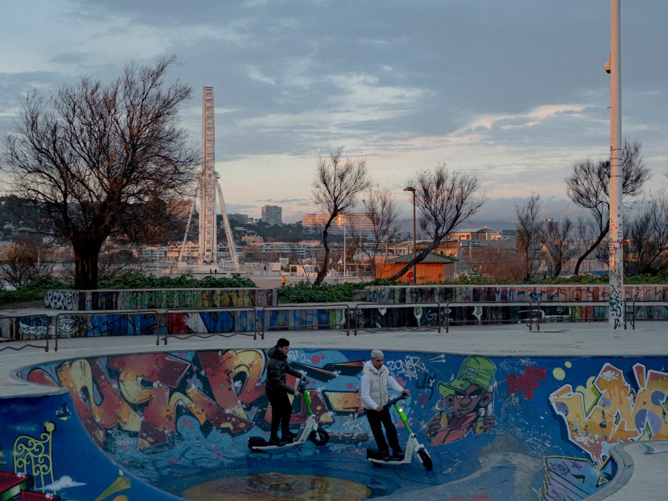 Two young men with scooters stand in a graffiti-covered concrete skate bowl at dusk in Marseille