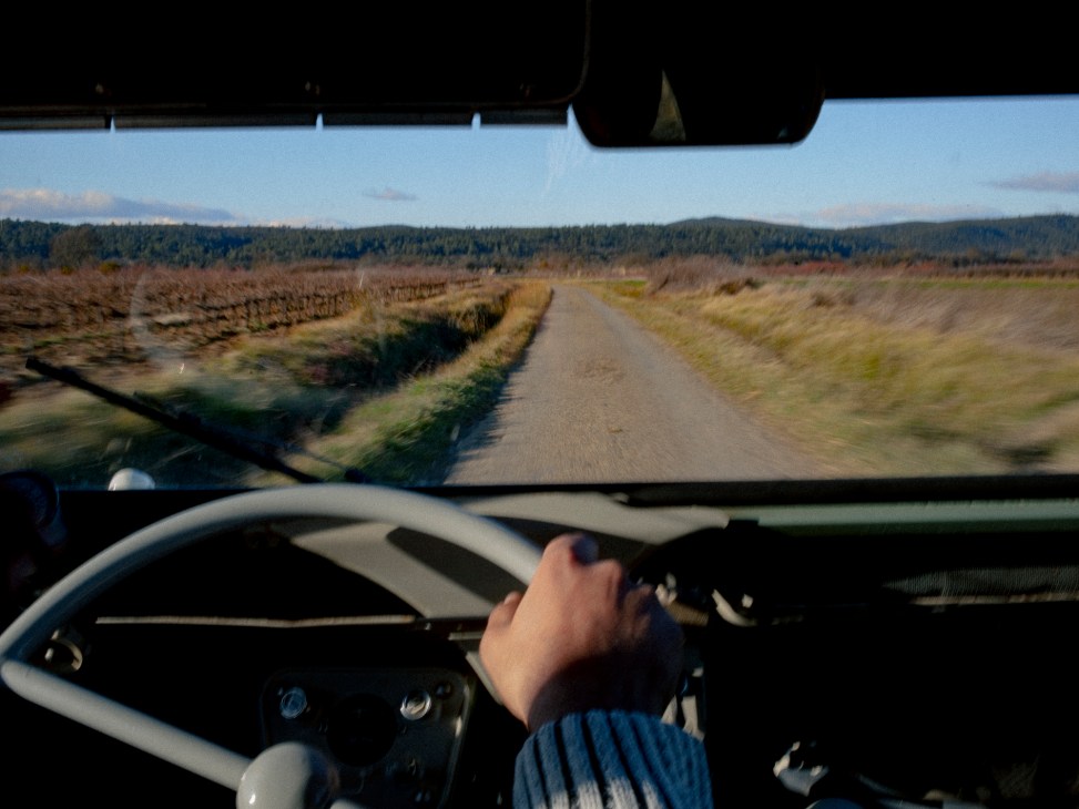 View from inside a car driving on a narrow rural road through countryside with rolling hills in the distance