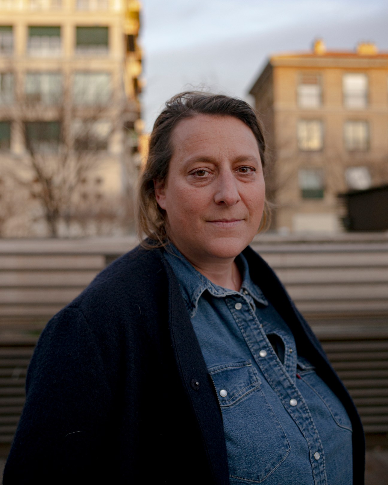 Middle-aged woman with dark hair wearing denim shirt and dark jacket standing outdoors in urban setting