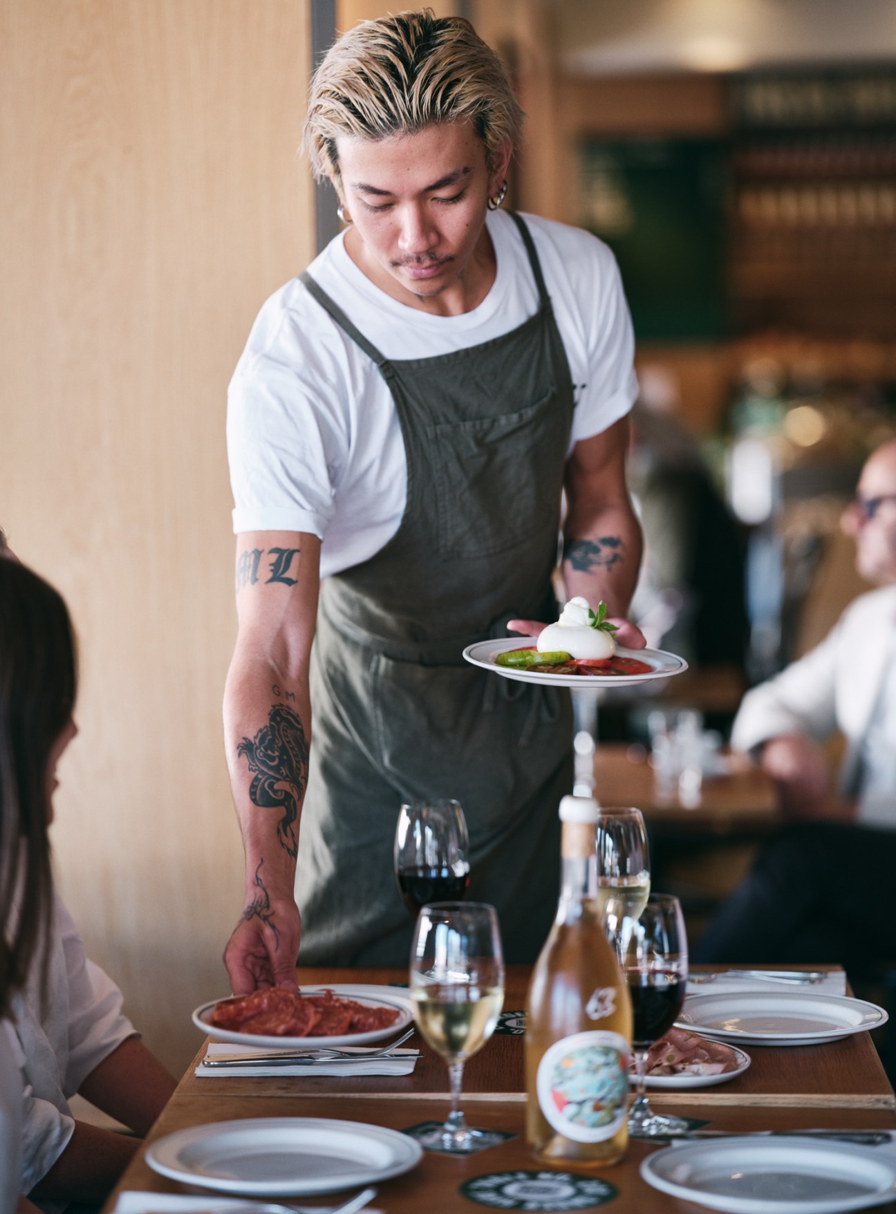 Tattooed chef with blonde hair serves plated dish in restaurant dining room