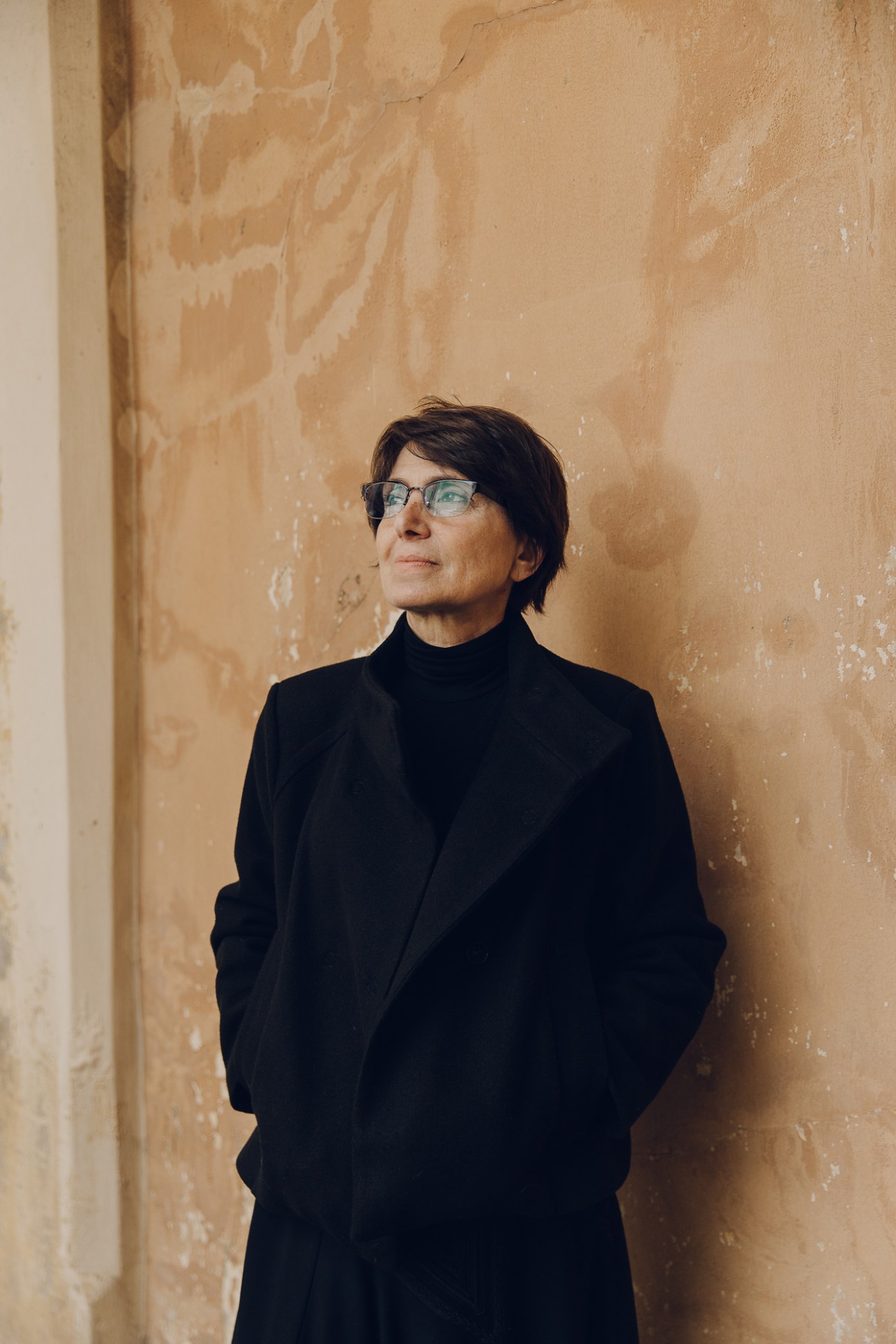 Annie Vartivarian standing against a weathered wall wearing glasses and a black coat