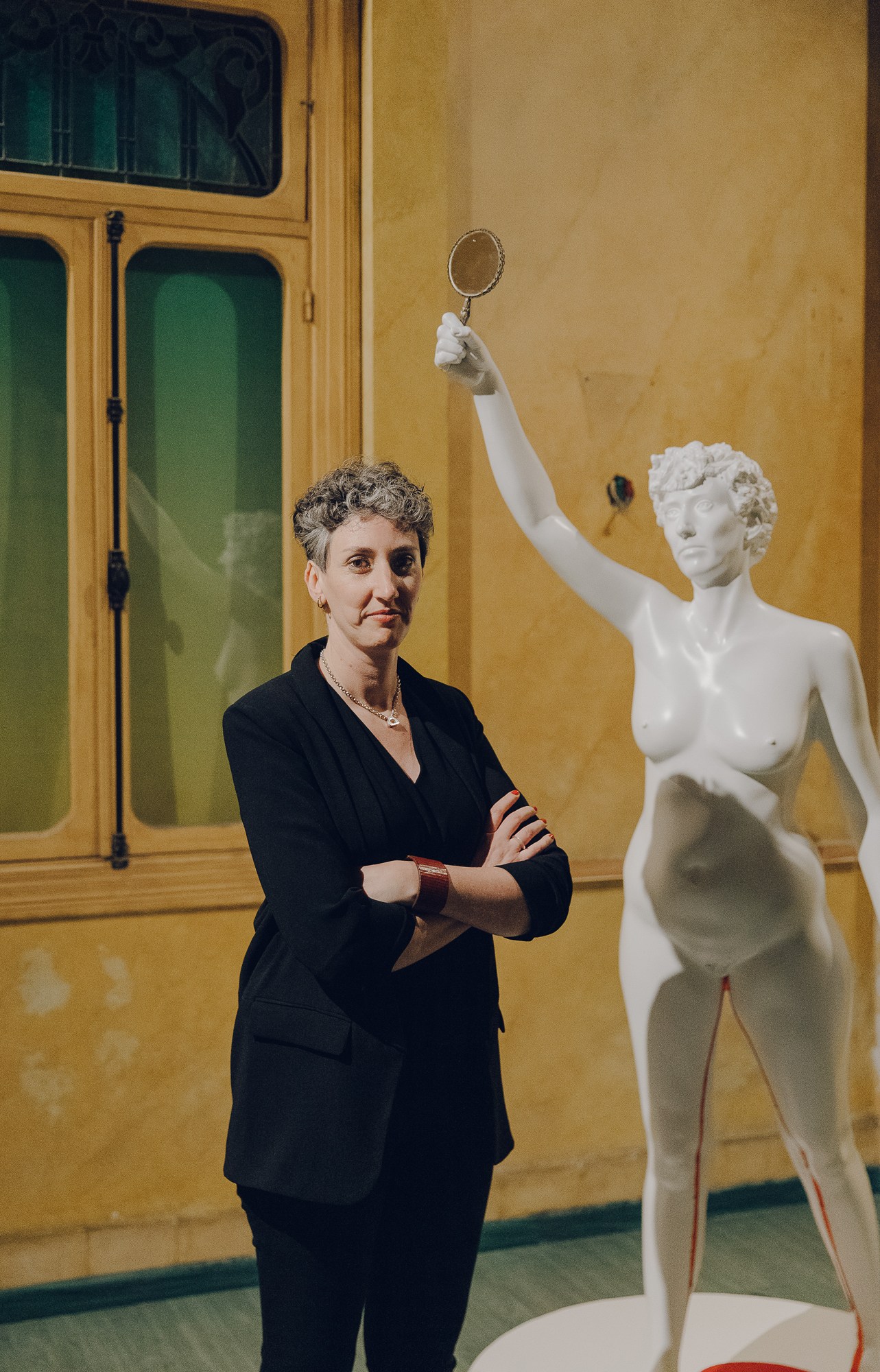 Visual artist Sirine Fattouh standing with arms crossed next to a white classical statue in a yellow gallery space