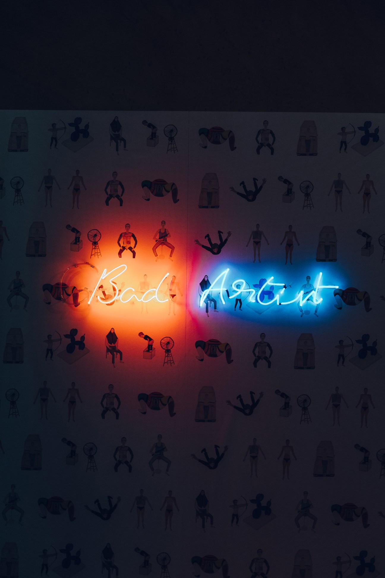 Neon sign reading 'Bad Artist' in cursive script illuminated against a dark background with silhouettes