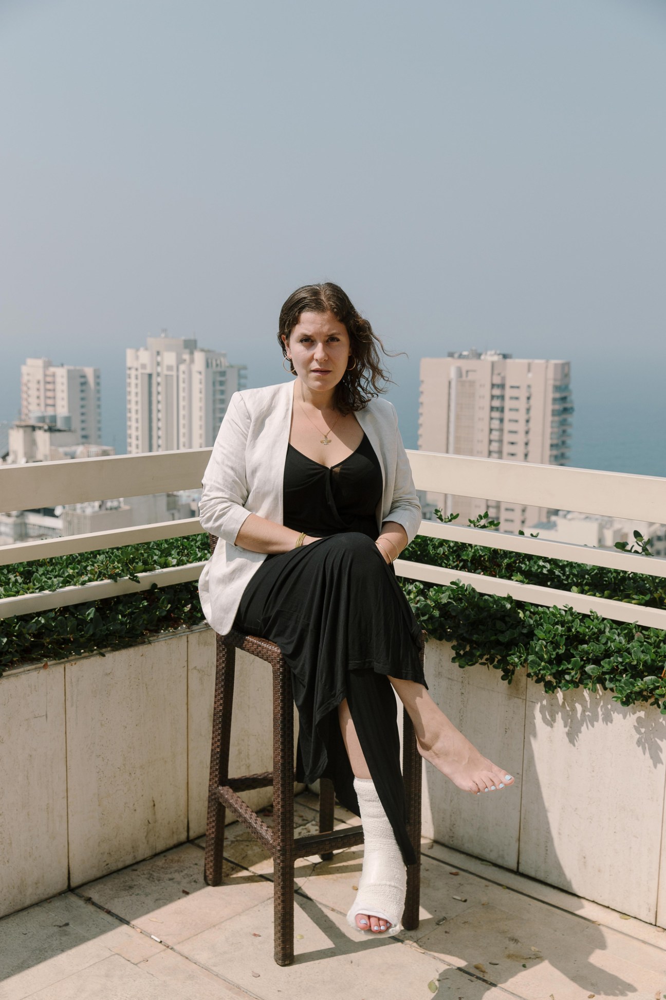 Woman in black dress and beige blazer sitting on balcony with city skyline in background