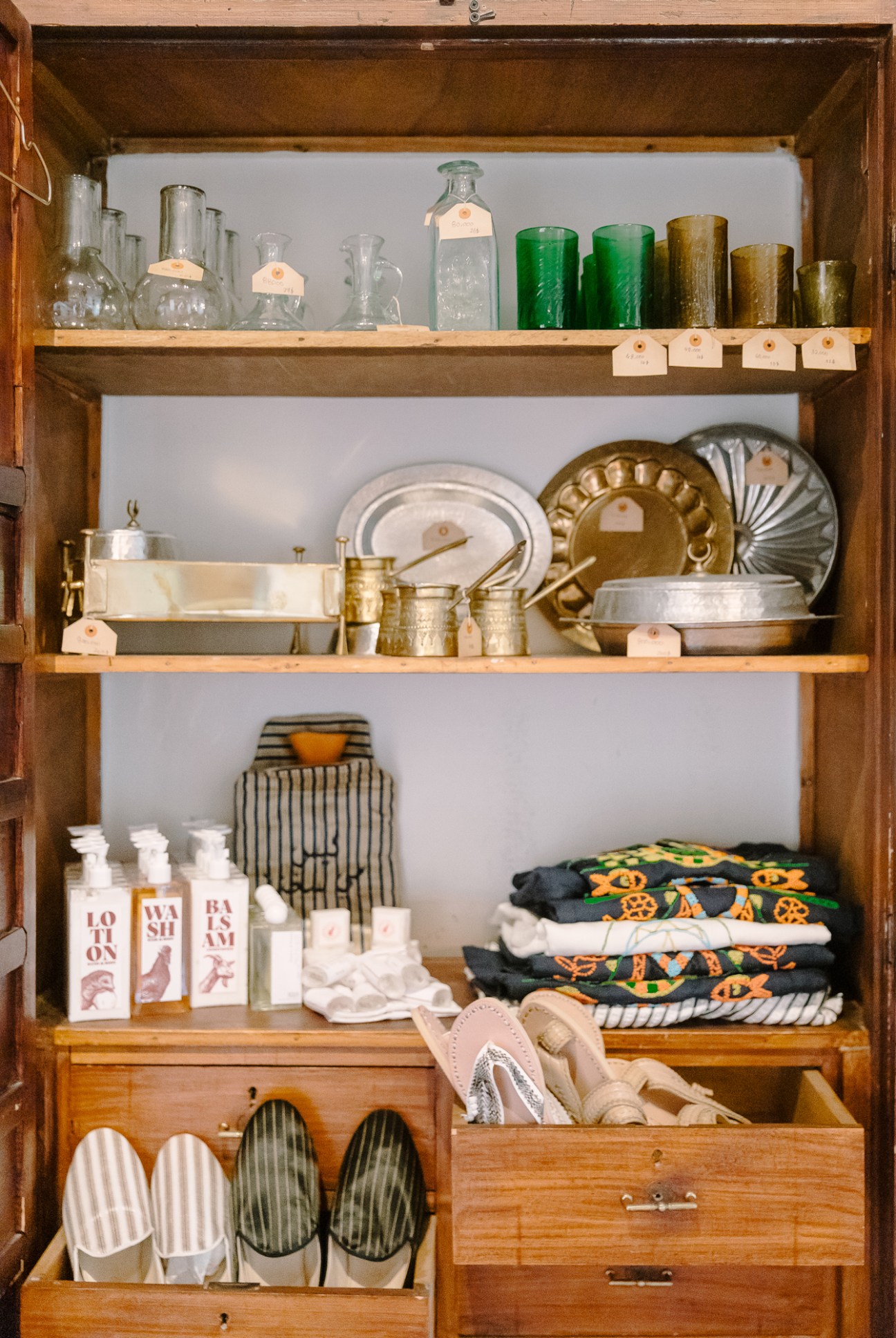 Wooden cabinet shelves displaying vintage glassware, brass vessels, decorative plates, bottled goods, textiles and household items.