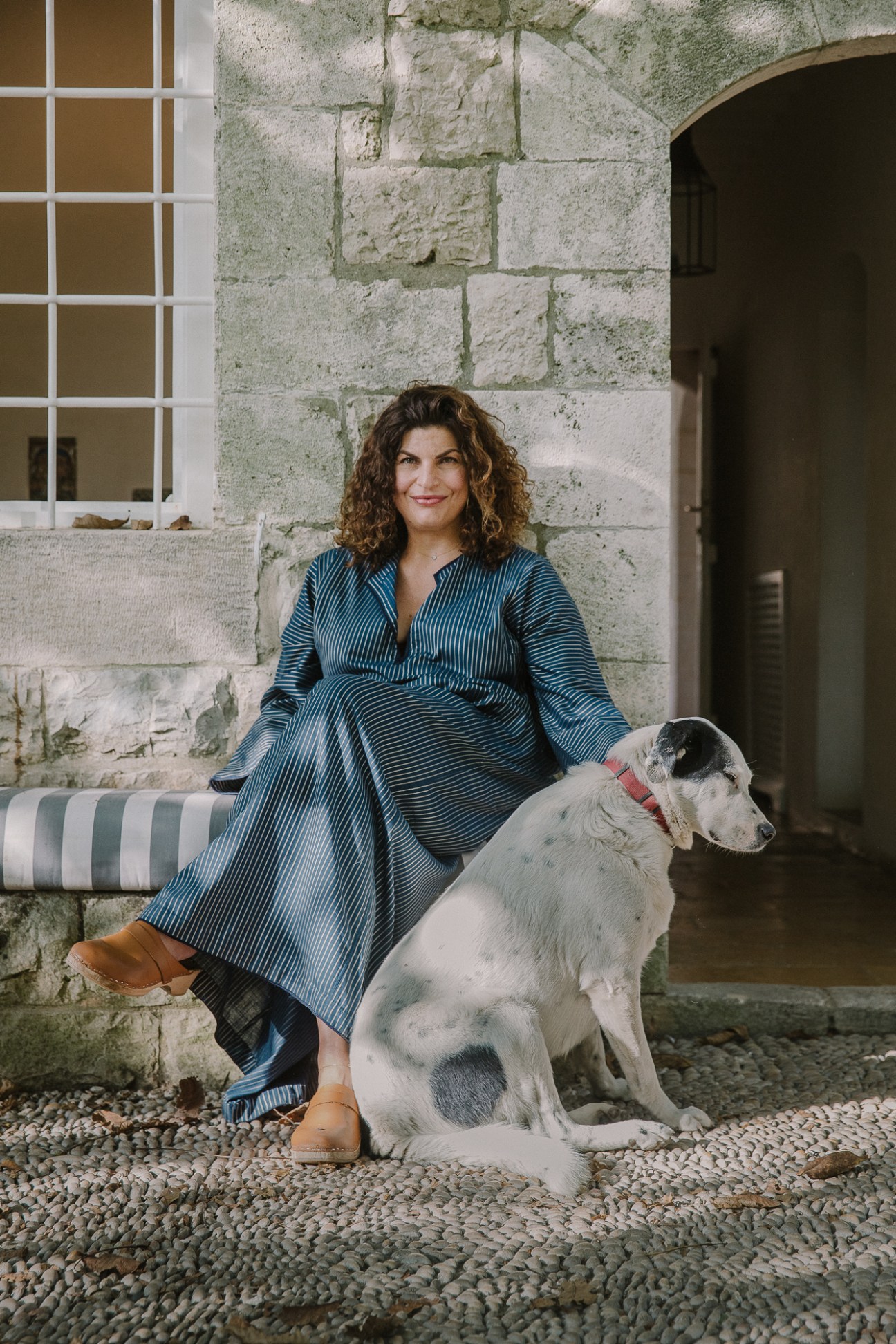 Woman in blue striped dress sitting against stone wall with white dog wearing pink collar