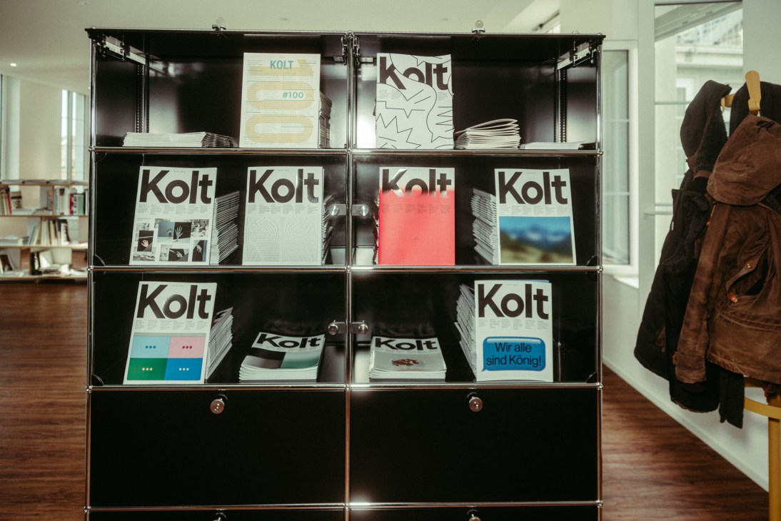 Modular shelving unit displaying multiple issues of Kolt magazine with various cover designs and colors.