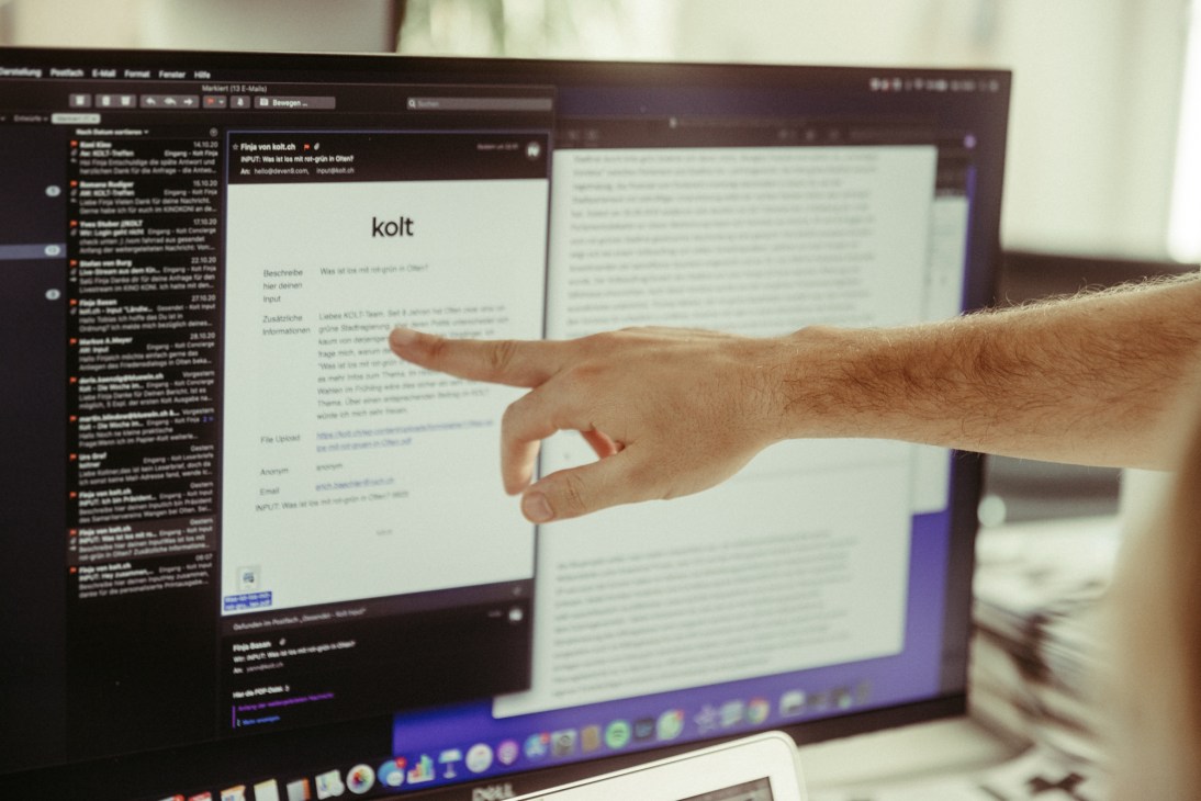 A hand points at a computer monitor displaying the Kolt website and digital platform interface.