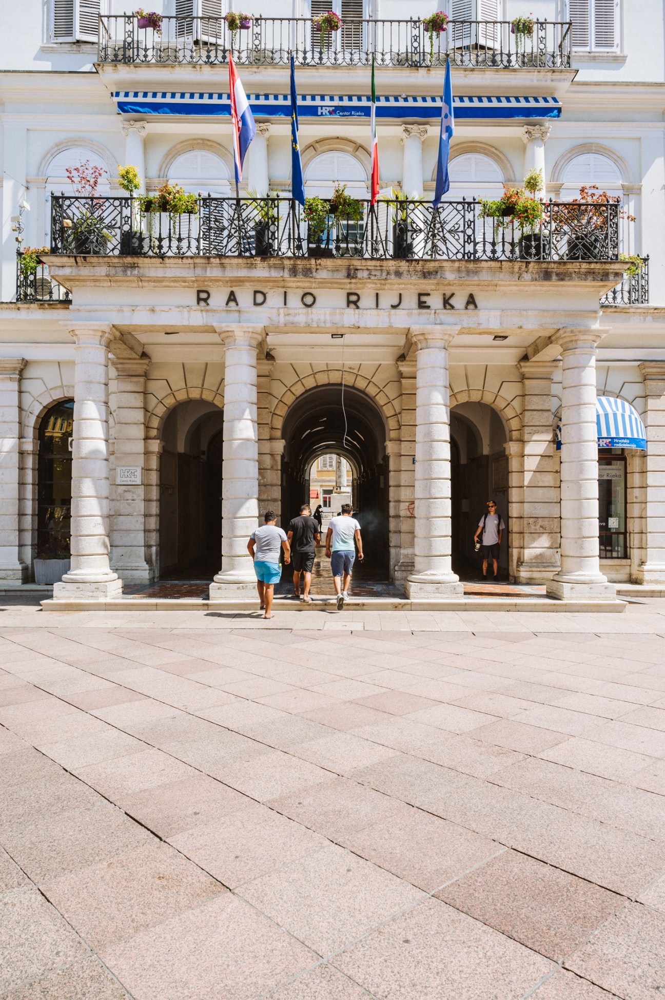 Historic stone building with arched entrances, ornate balconies, and flags, with people walking through the archways