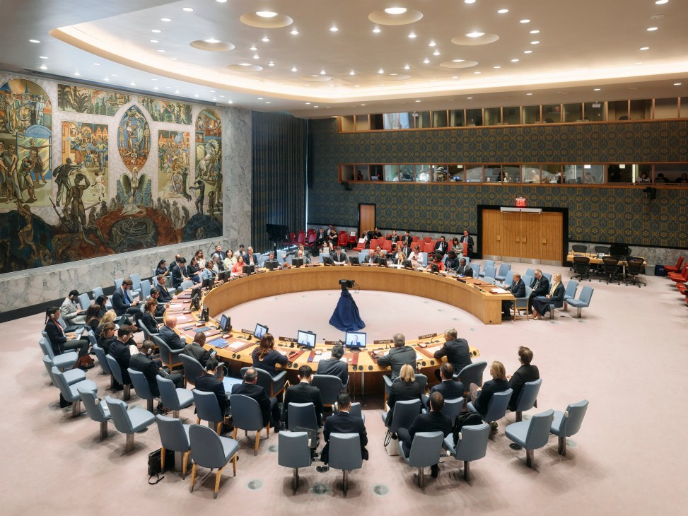 UN ambassadors seated around a curved wooden table in the Security Council chamber during a session.