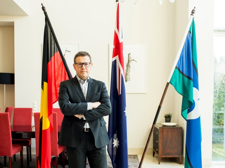 Paul Wojciechowski, Australia's ambassador to Poland, stands with arms crossed beneath a white chandelier, flanked by three flags.