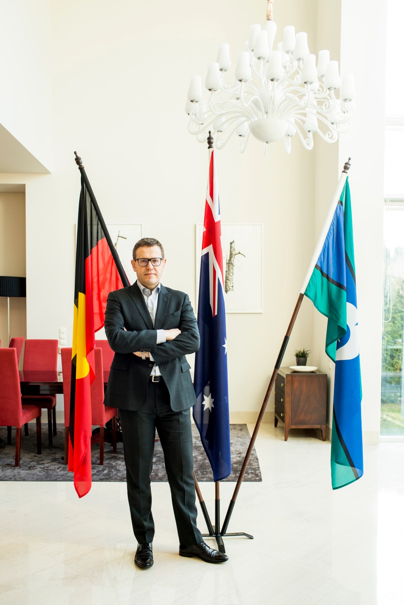 Paul Wojciechowski, Australia's ambassador to Poland, stands with arms crossed beneath a white chandelier, flanked by three flags.