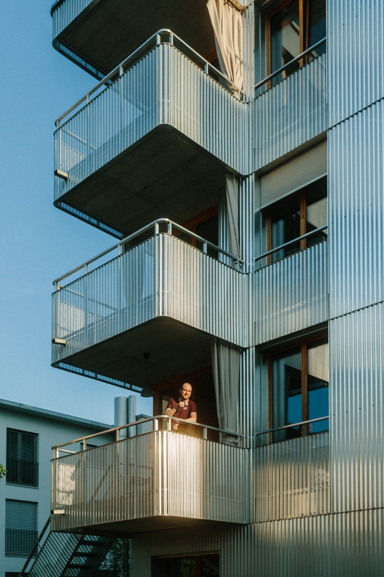 Resident standing on a balcony of Küsnacht apartments with anodised aluminium railings and corrugated metal cladding.
