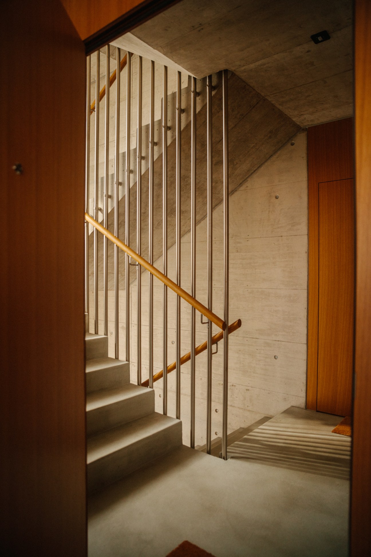 Interior staircase with wooden handrail, white vertical slats, concrete walls and orange door at Küsnacht apartments.