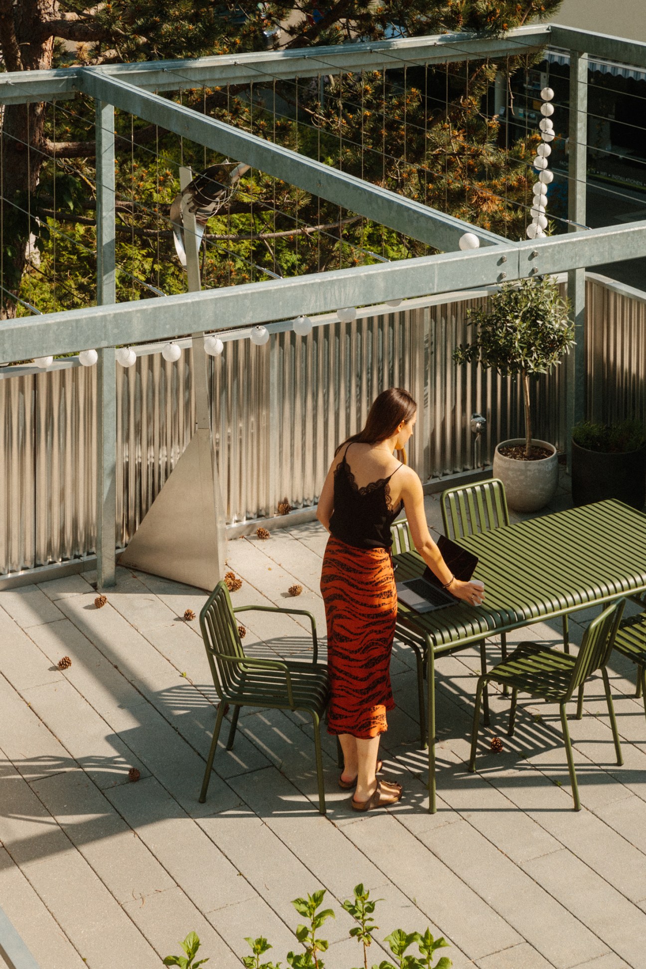 Woman standing on a modern apartment building terrace with metal railings, green chairs, and potted plants overlooking green vegetation.