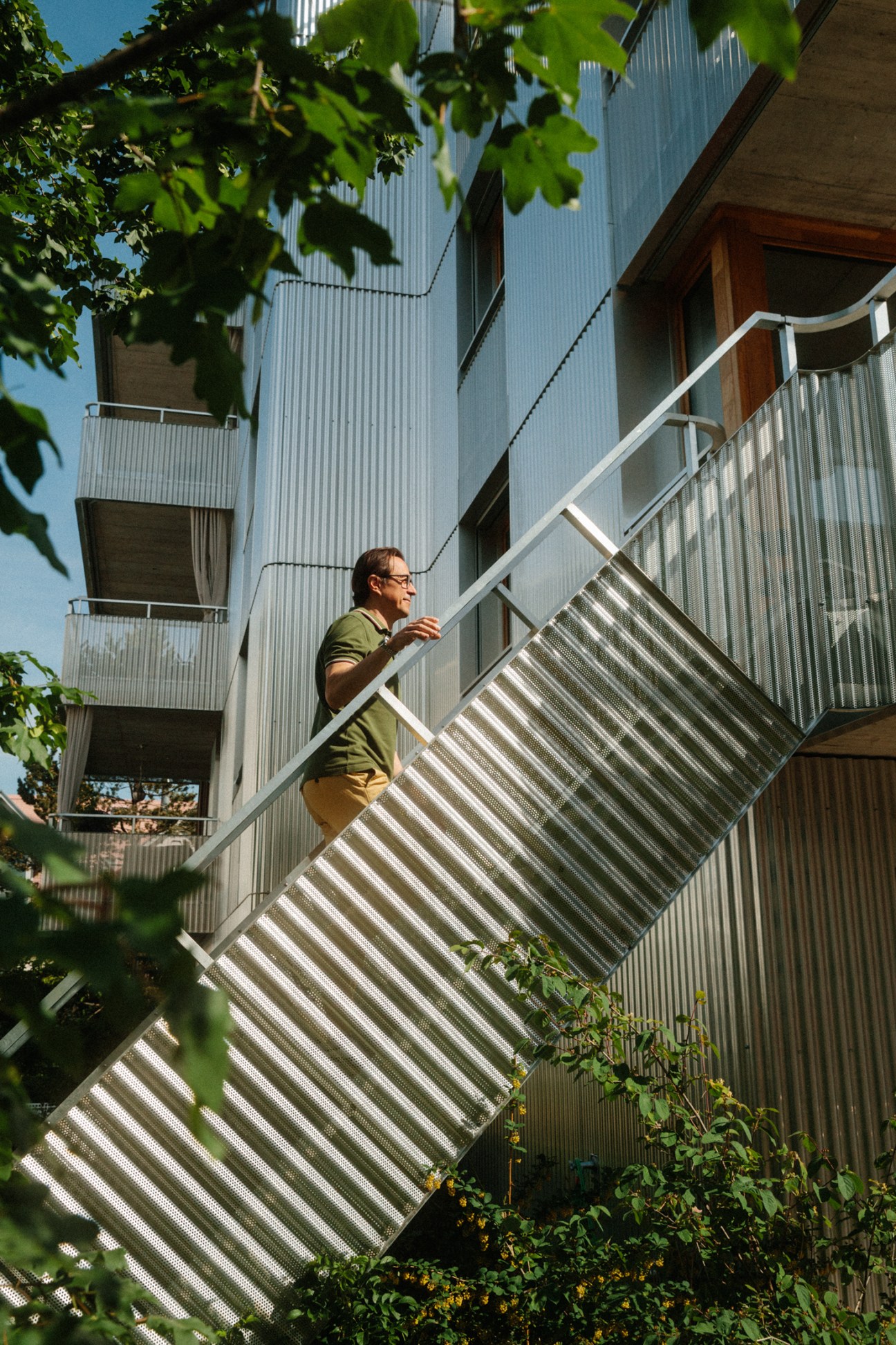 Man in olive shirt climbing metal staircase on Küsnacht apartments building with corrugated metal facade.