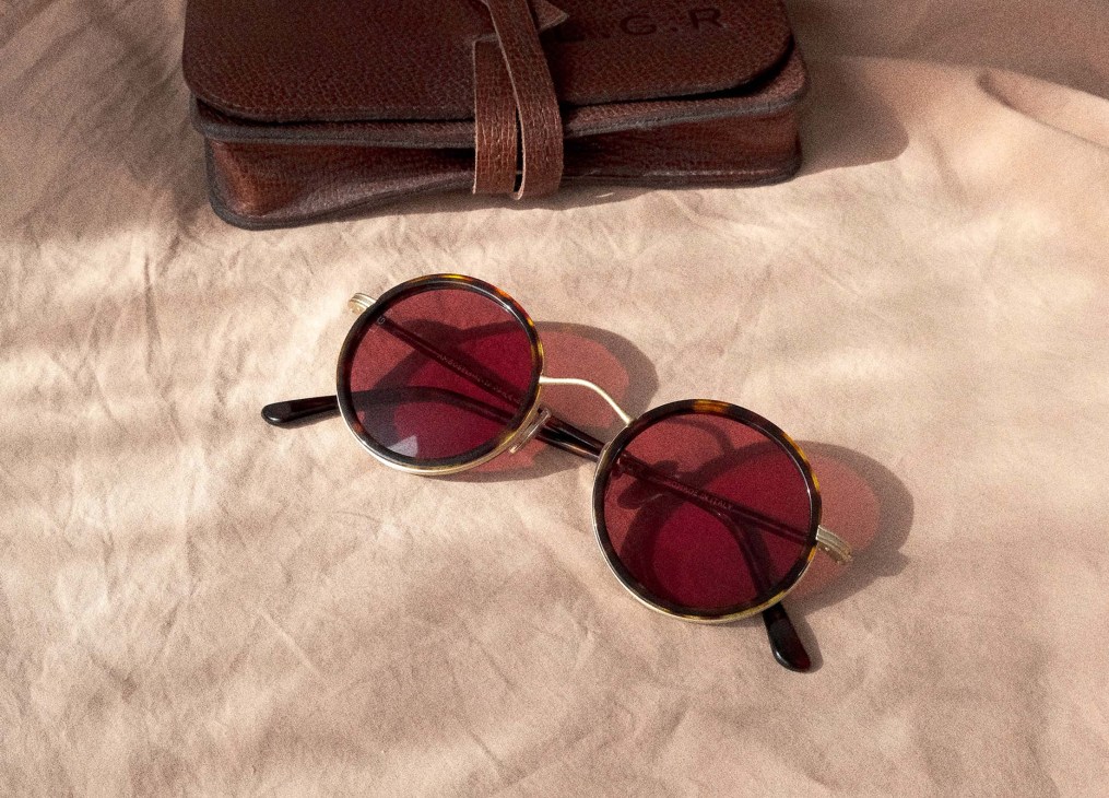 Pair of brown tortoiseshell-frame sunglasses with red-tinted lenses and a brown leather case on a beige fabric surface.