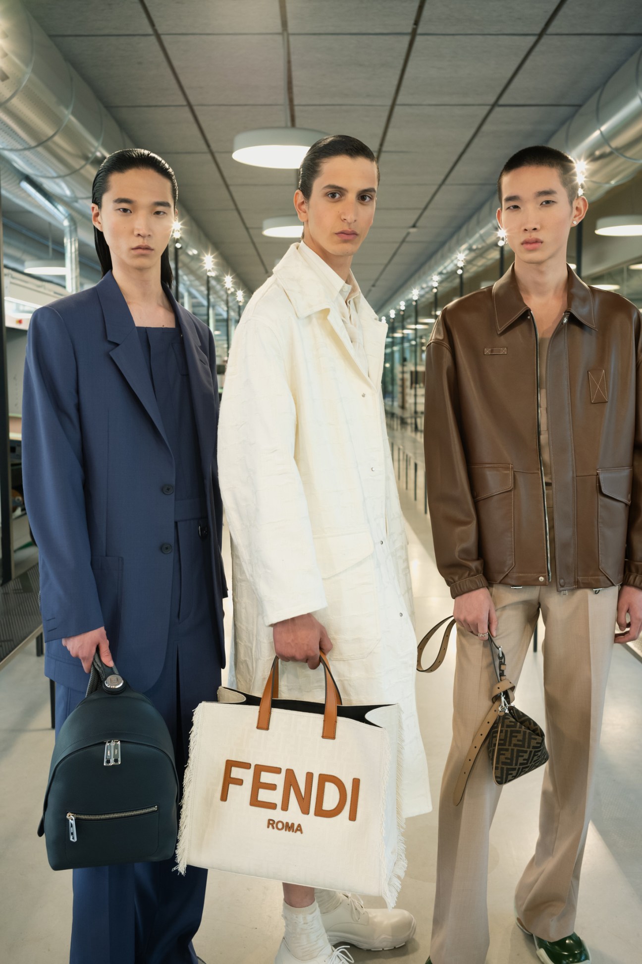 Three male models in tailored outerwear stand in a modern office hallway, one holding a white Fendi shopping bag