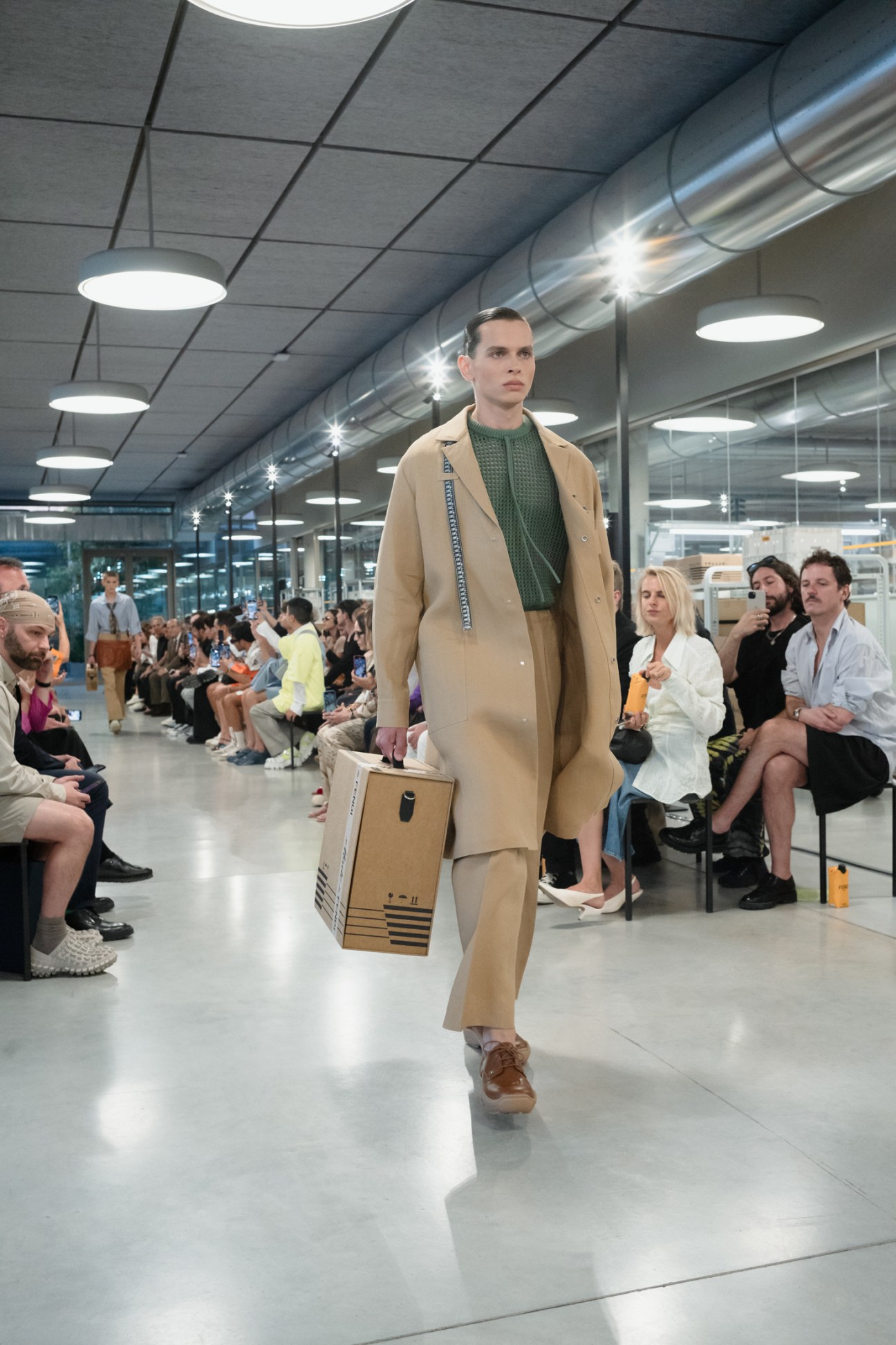 Male model walks runway in beige coat and green sweater carrying cardboard box at fashion show in modern venue