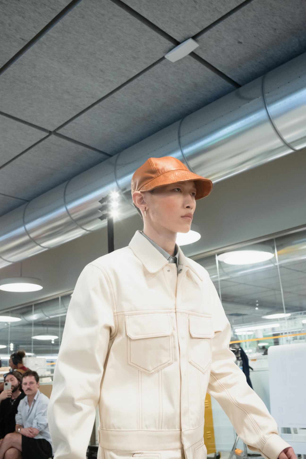 Model wearing cream workwear-inspired jacket and orange leather cap in modern industrial setting