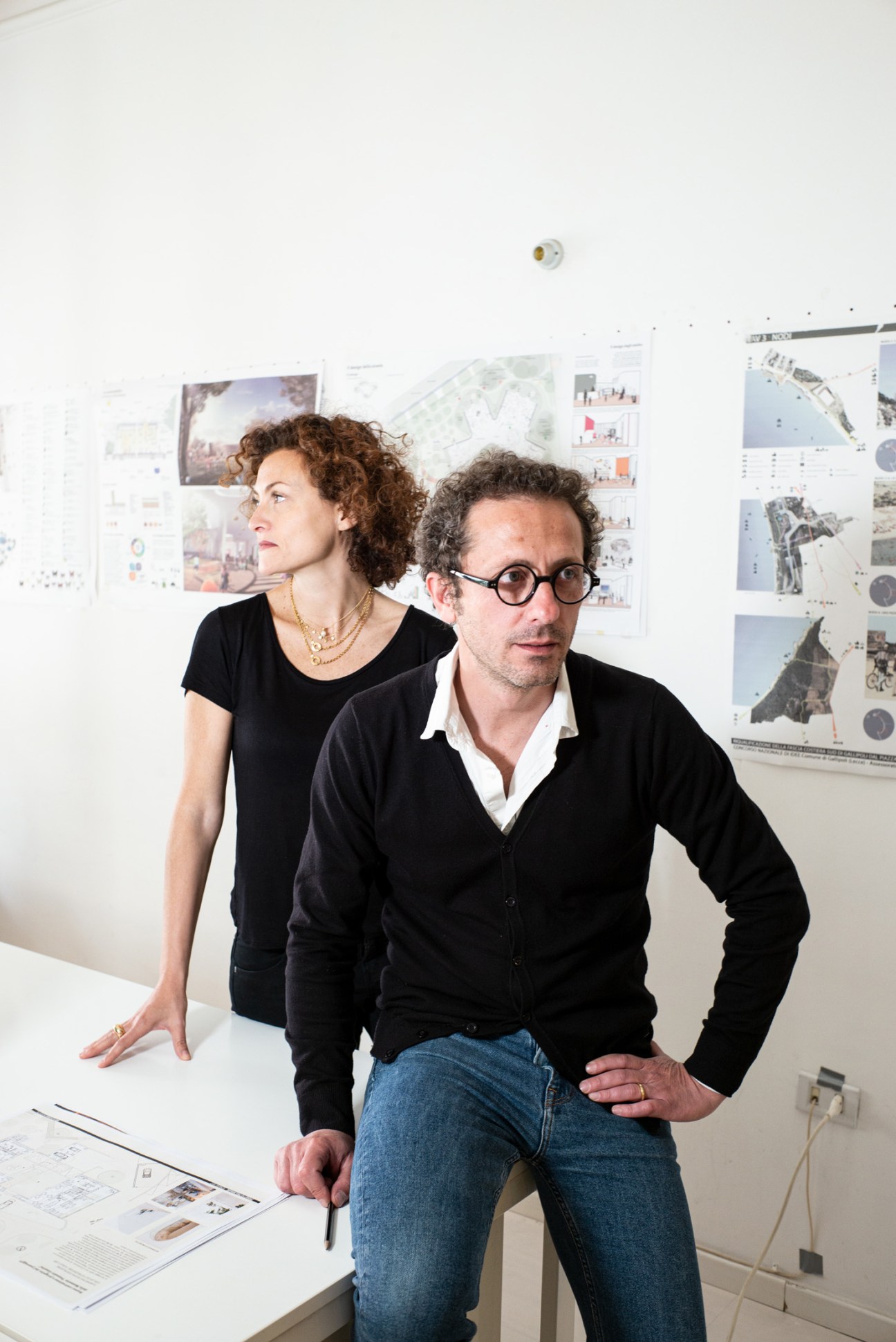 Two architects stand in an office with design plans and drawings pinned on the wall behind them.