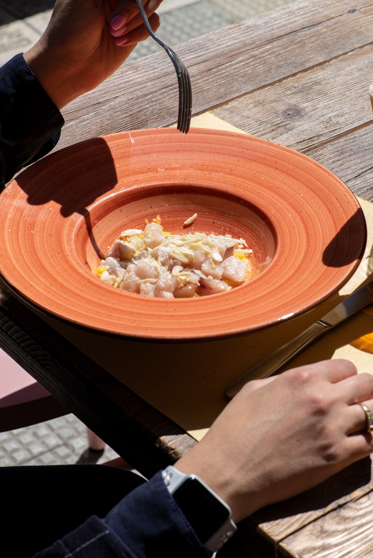A hands holding a fork over an orange ceramic plate of fresh fish at Al Coltello restaurant in Bari.