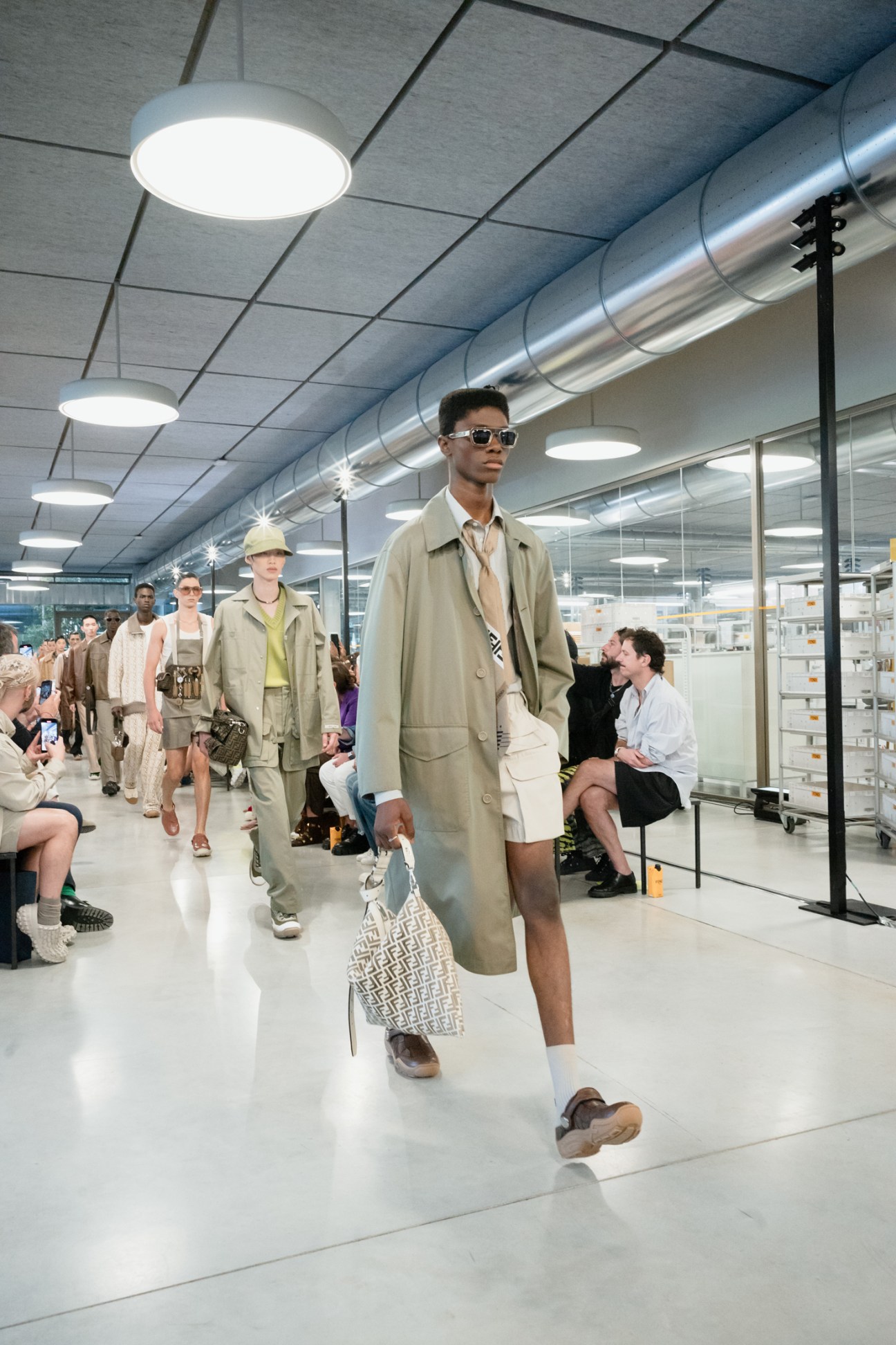 Male model in beige trench coat and sunglasses walks down runway in industrial-style venue with seated audience