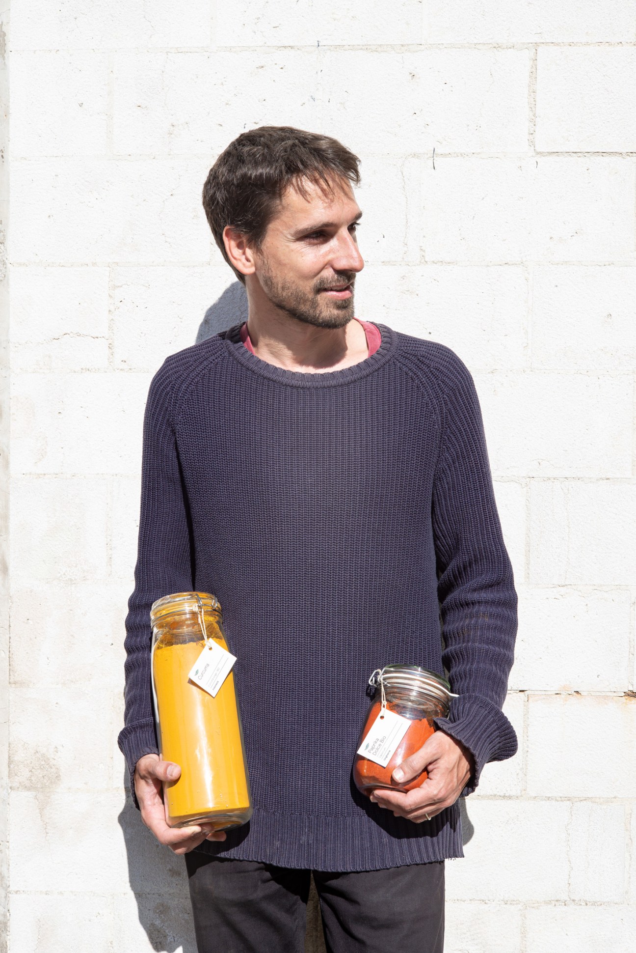 Man holding glass jars of yellow and orange preserves against a white brick wall.