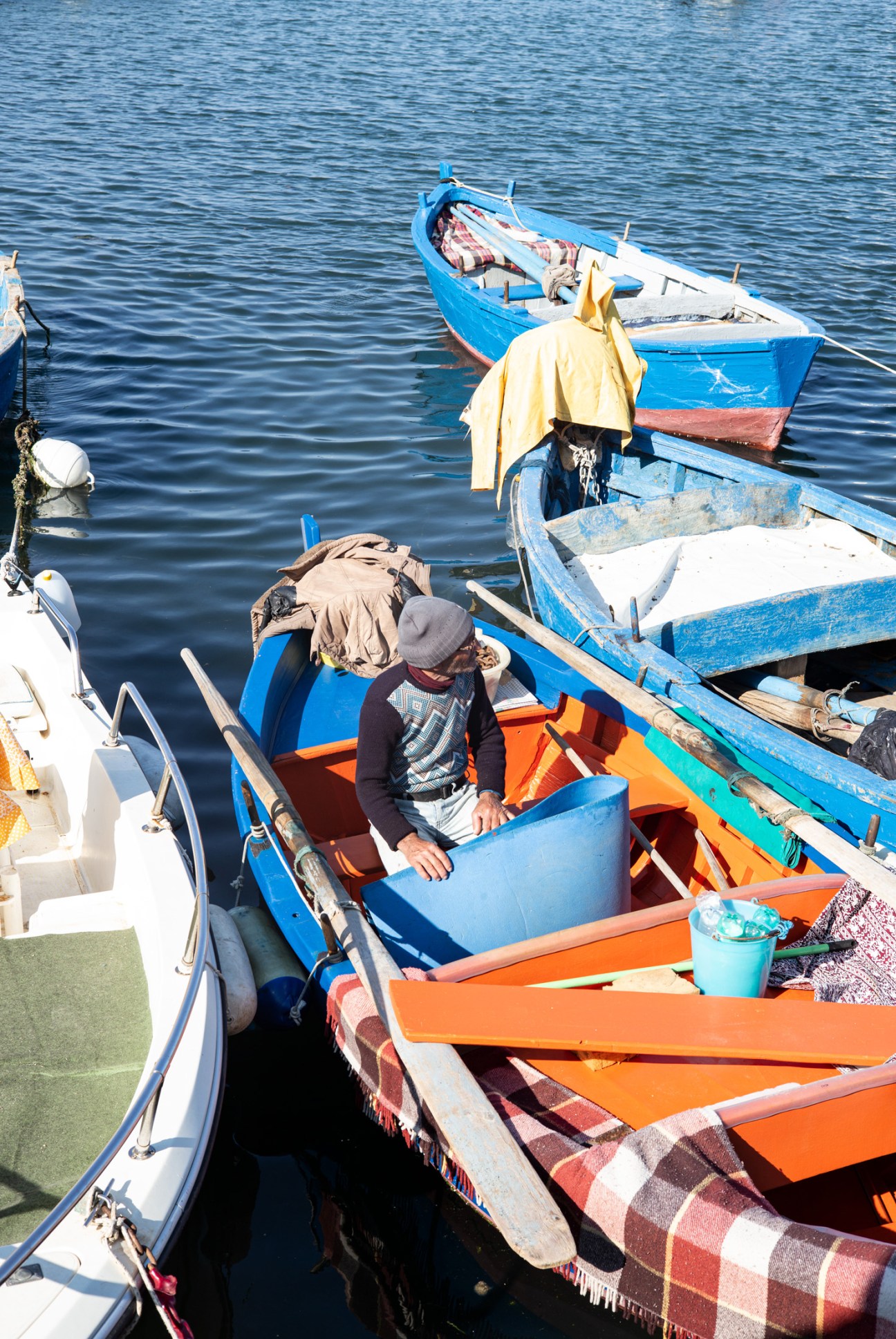 A fisherman sits in a blue and orange boat moored in calm Mediterranean waters near Bari, Italy.