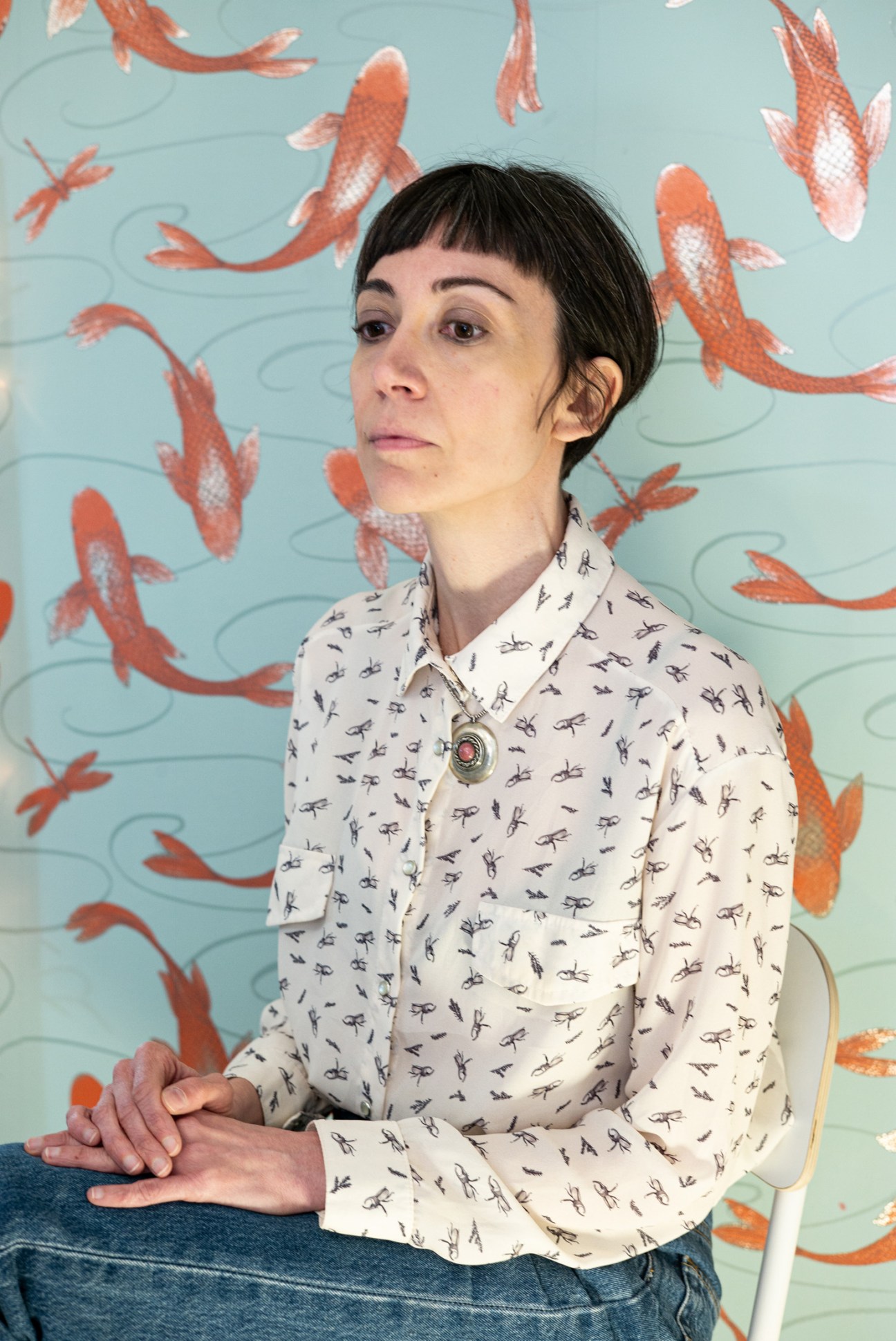 Woman with short dark hair wearing a cream shirt with dragonfly print, posing against a turquoise wall decorated with red koi fish illustrations.