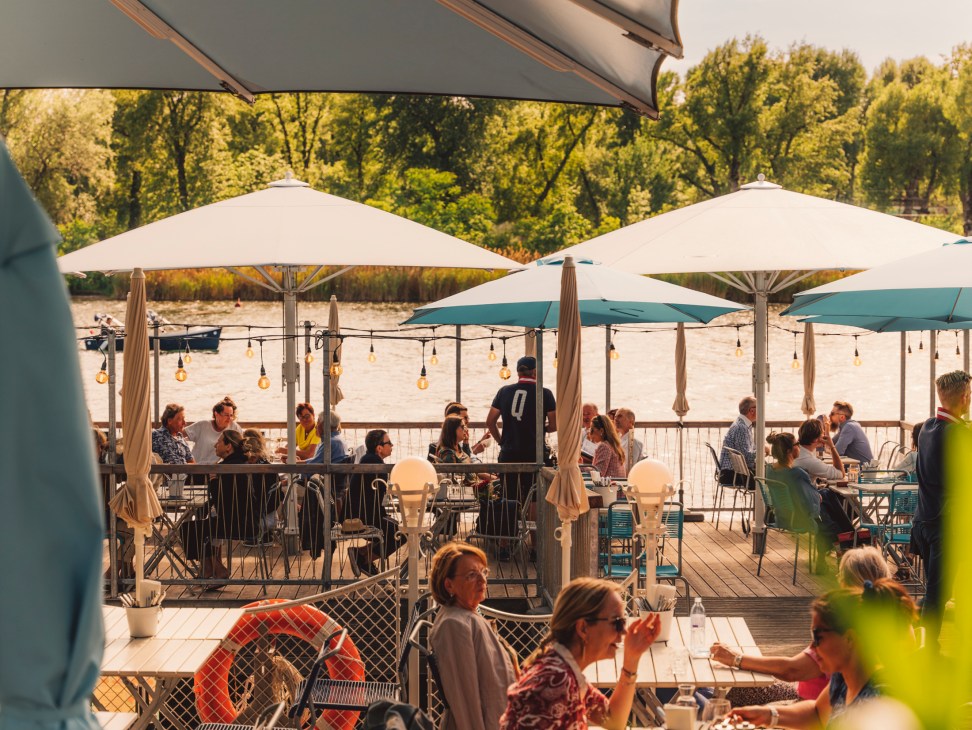 Waterfront restaurant terrace with diners under umbrellas overlooking harbor with boats and trees
