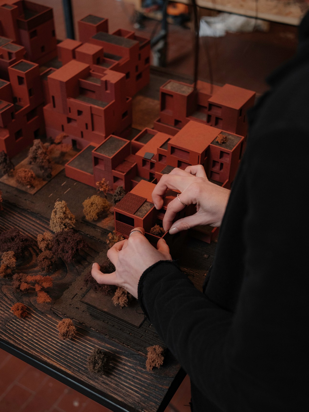 Hands positioning miniature architectural model buildings on a wooden surface with landscaping materials