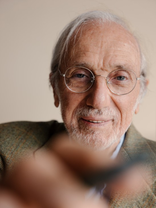 Portrait of Renzo Piano in his Genoa studio, sitting at a table with architectural drawings and models, with large windows overlooking the Ligurian Sea