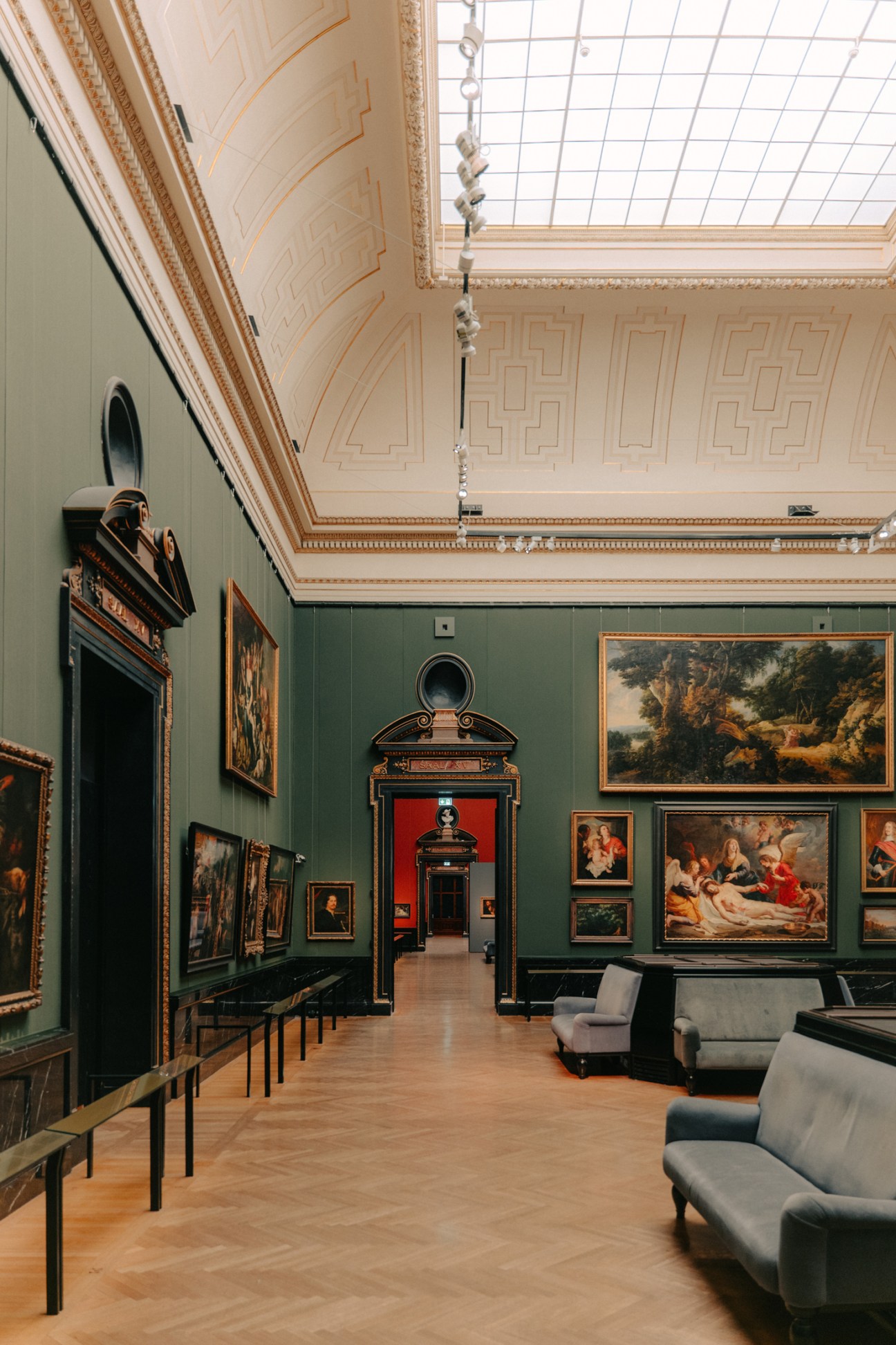 Interior of a grand gallery room with green walls displaying classical paintings in ornate frames, decorated ceiling with geometric patterns, and upholstered seating