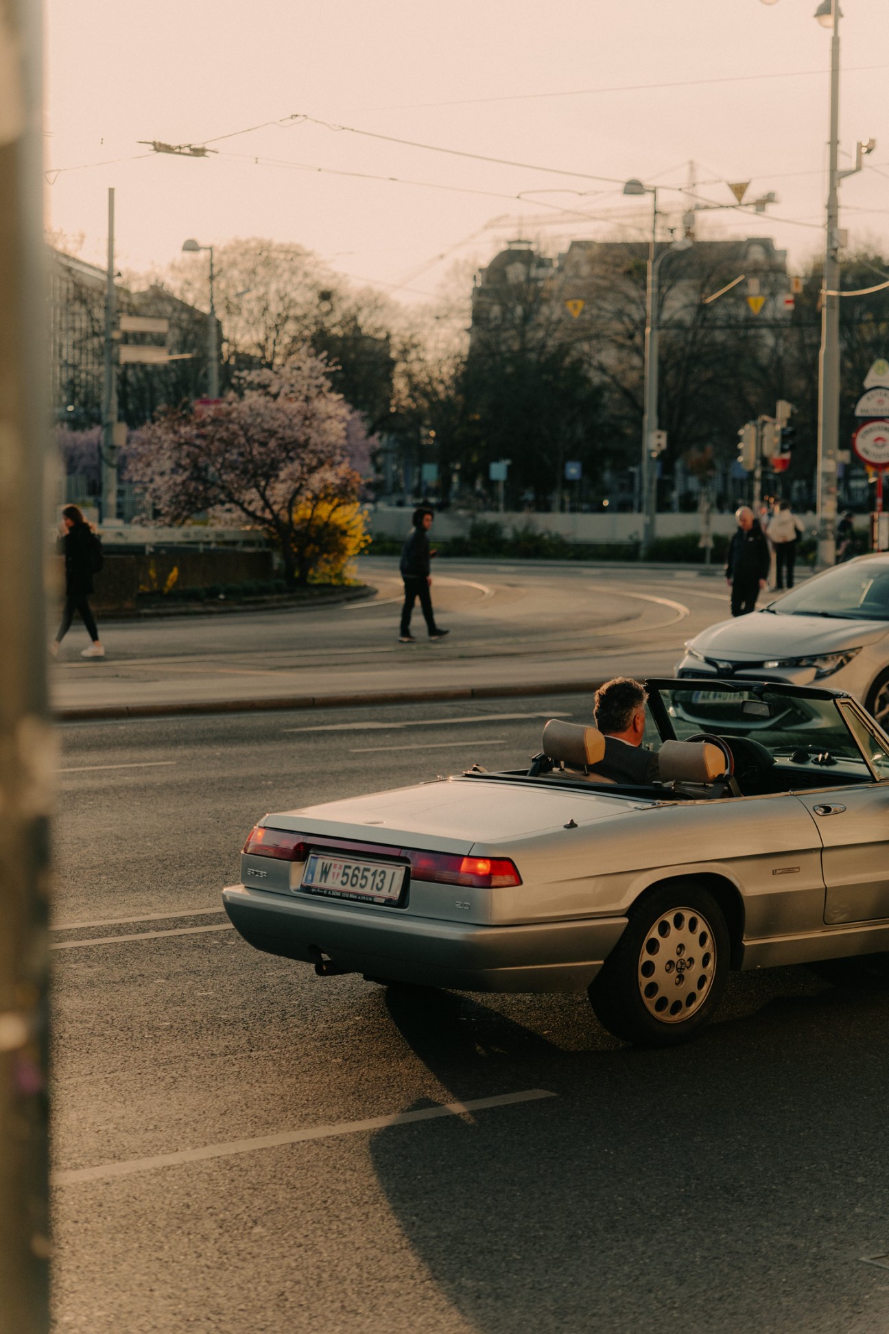 A convertible car with driver stopped at a Vienna intersection, with pedestrians and bare trees in the background