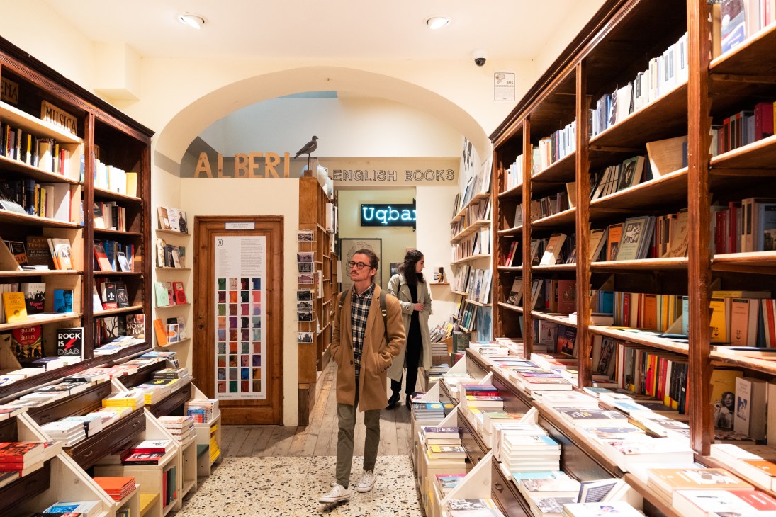 Man in glasses and tan coat browsing books in wooden-shelved bookstore with arched ceiling