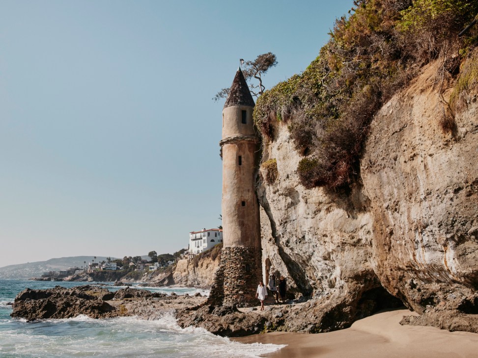 Medieval-style stone tower with conical roof built into coastal cliff face above rocky shoreline and ocean waves