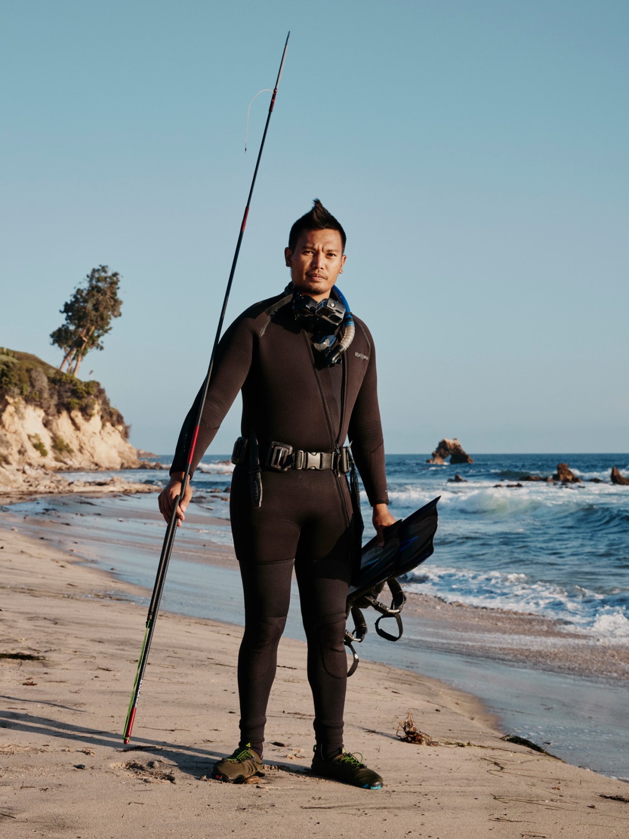 Person in wetsuit holding spearfishing equipment standing on beach with ocean waves in background