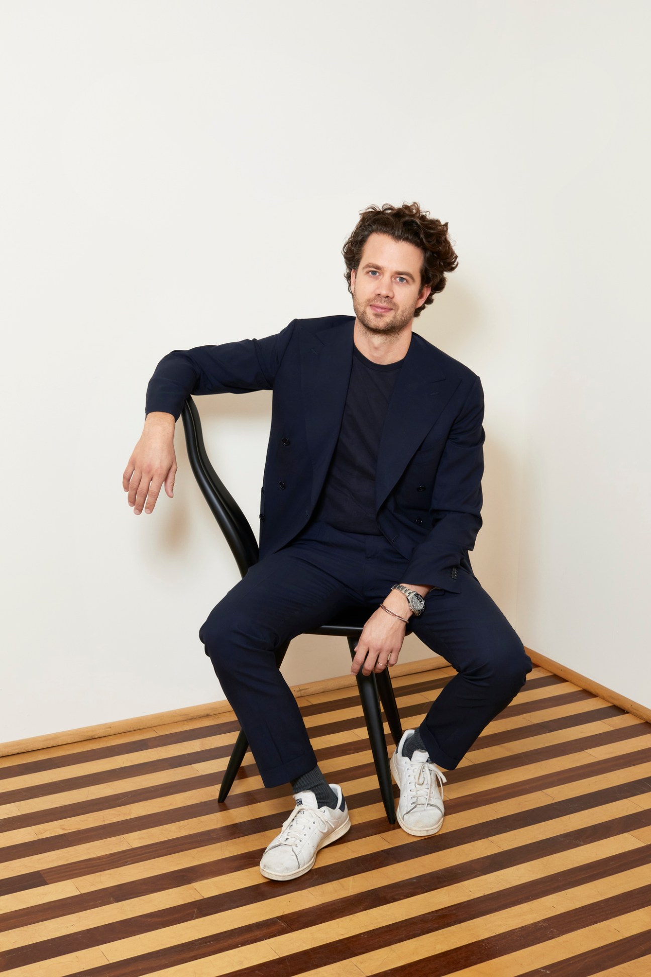 Man with curly hair in navy suit sitting casually on black chair against white wall