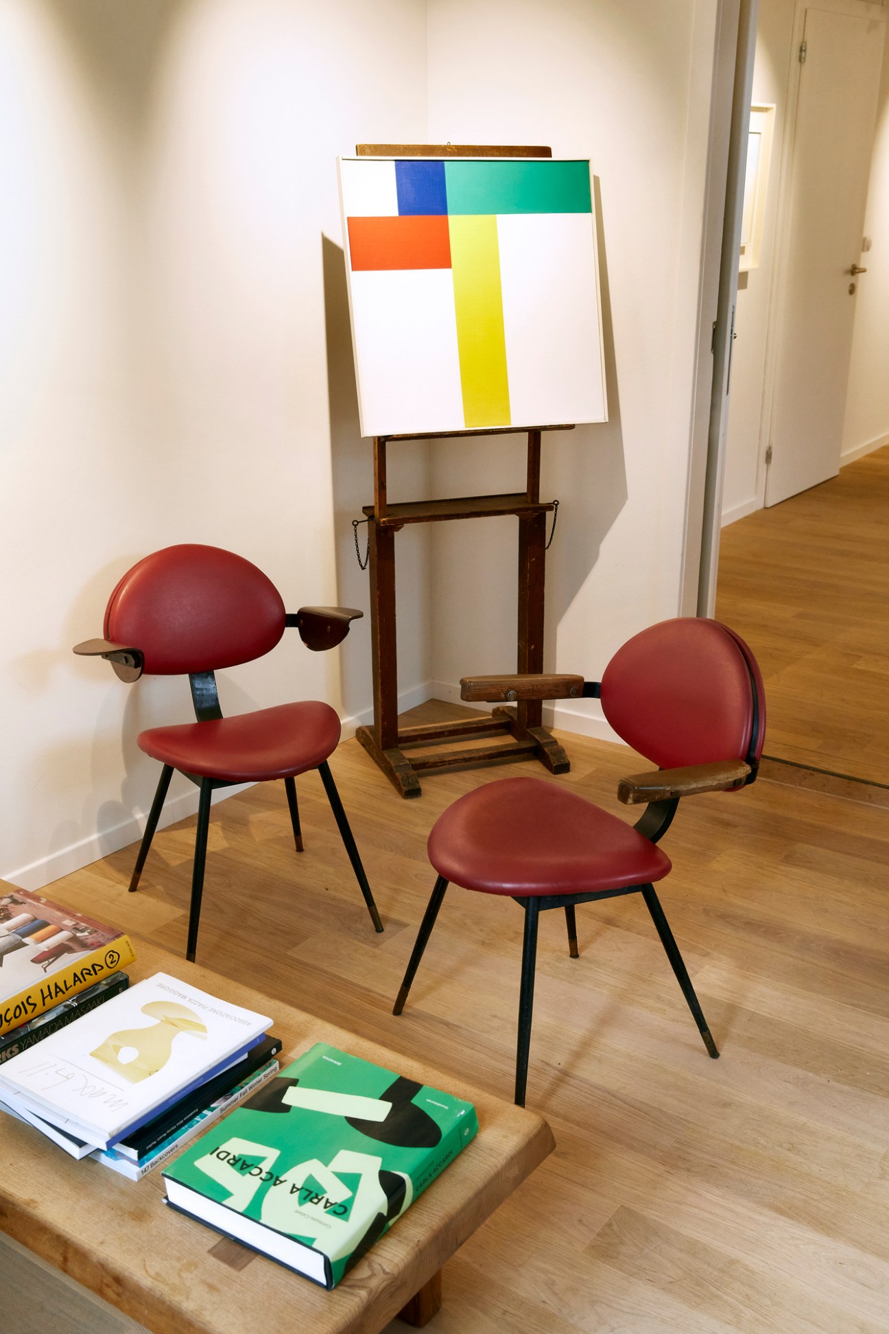 Abstract geometric painting with colorful rectangles on easel beside red chairs and art books