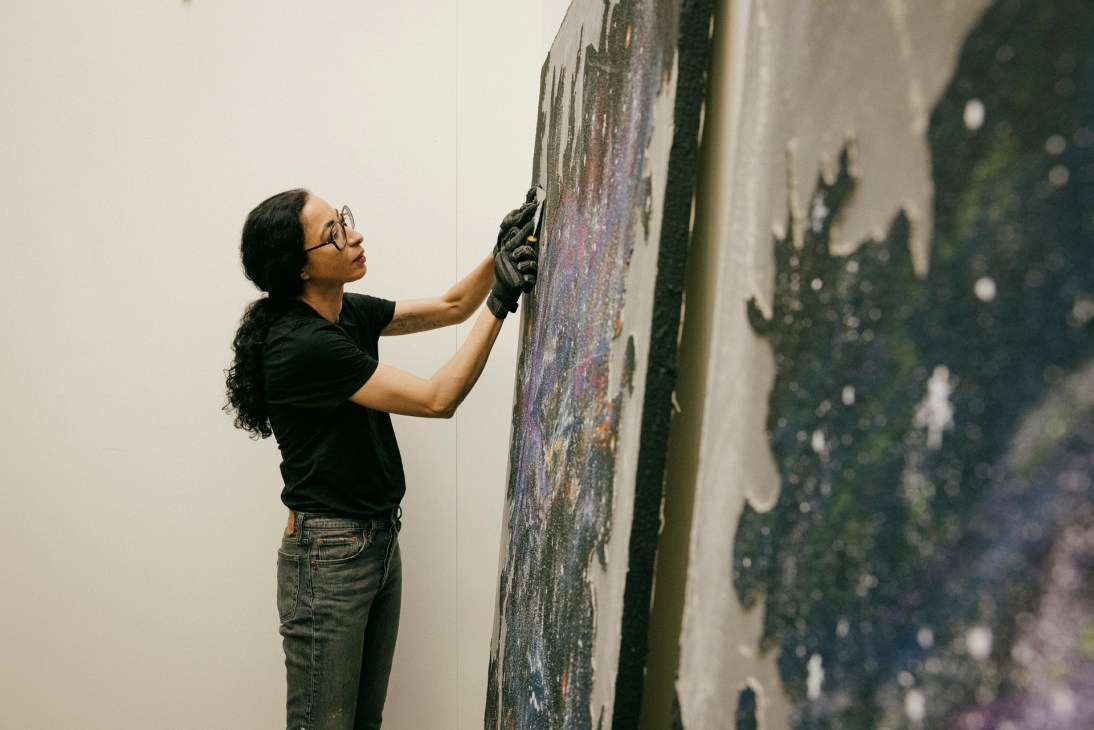 Artist in black shirt and gloves working on a large colorful painting on a white studio wall