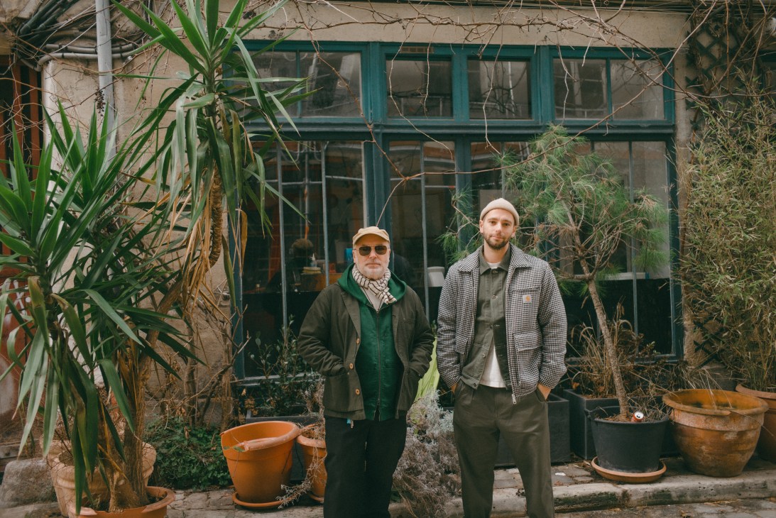 Two men standing outside with potted plants, one wearing a beanie and jacket, the other in checkered coat