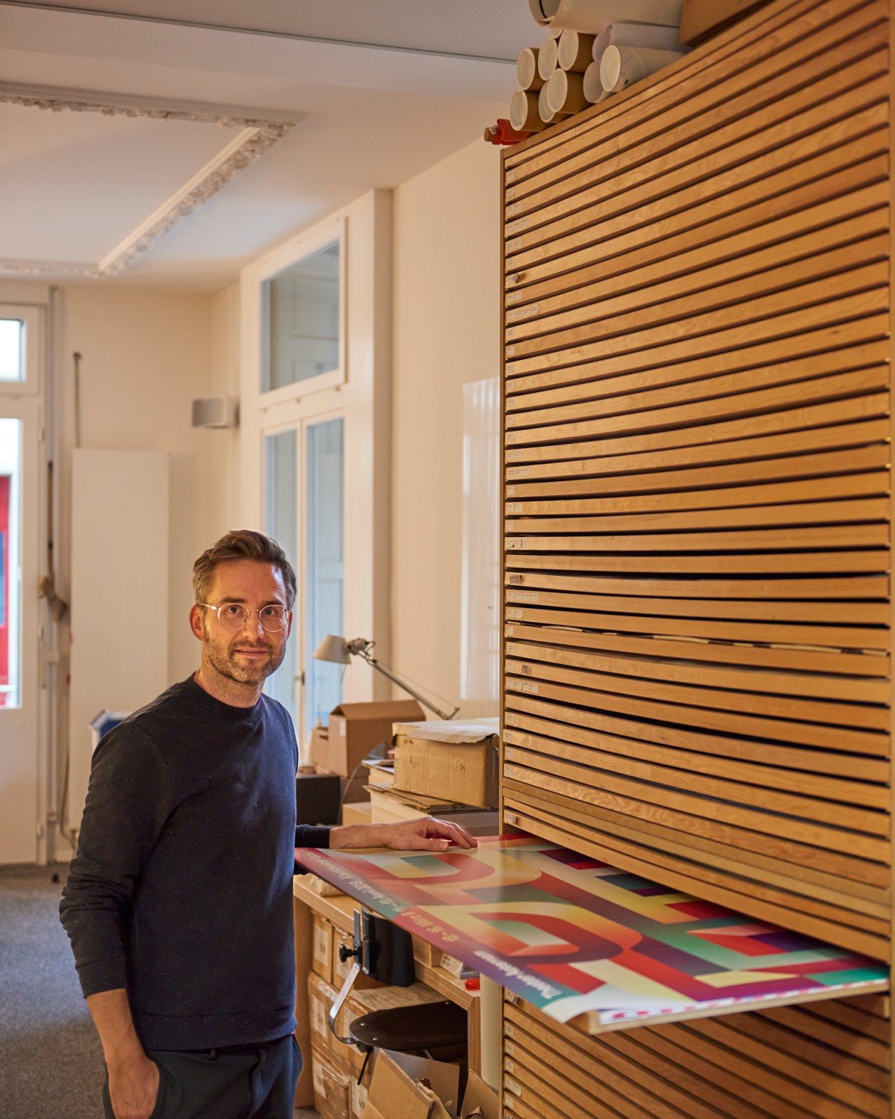 Man with glasses and dark sweater standing at wooden desk with colorful poster designs and wooden slat wall behind