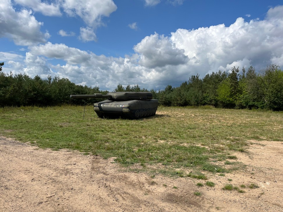 Inflatable tank decoy in an open field with forest and cloudy sky in background.