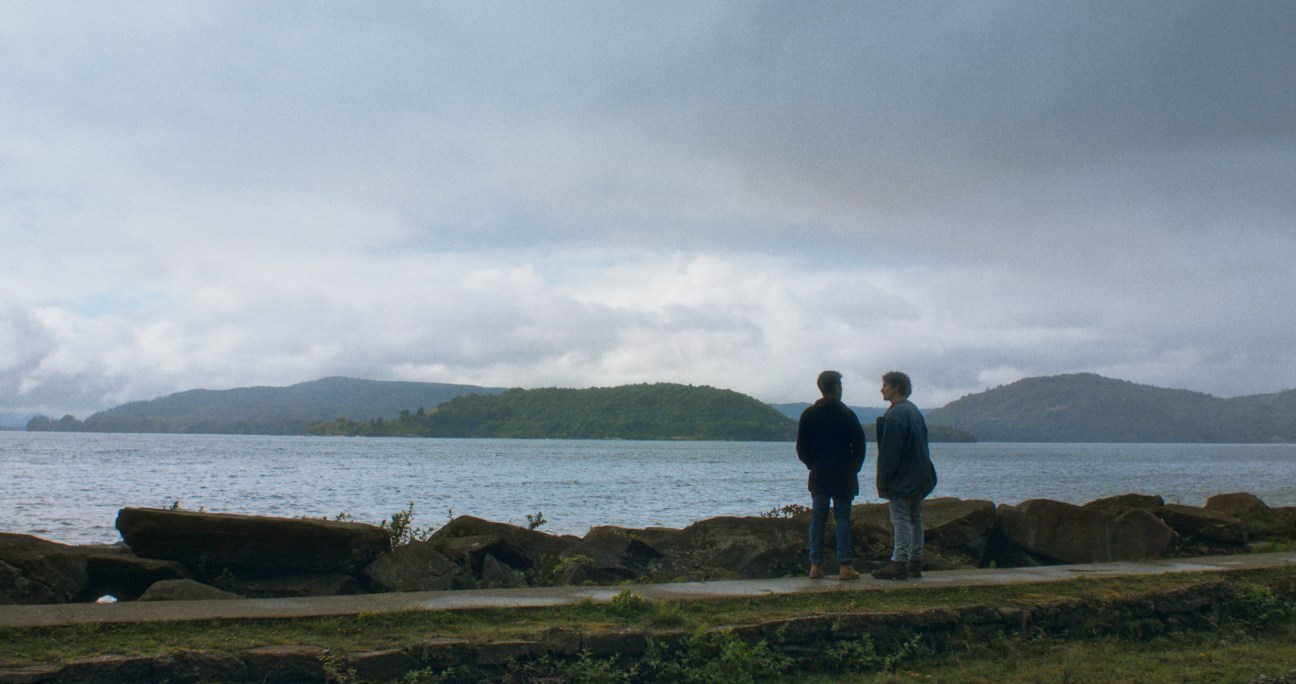 Two men standing by a lake looking at forested hills in the distance under an overcast sky