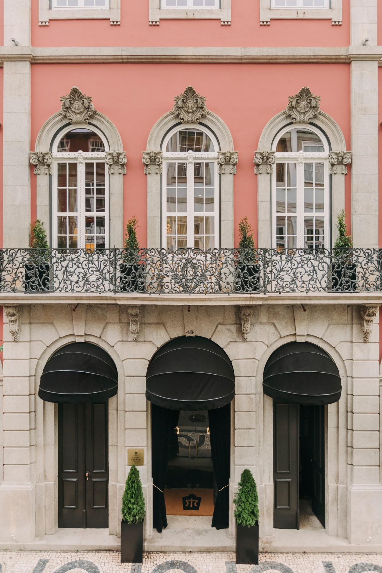 Pink historic building facade with three ornate arched French doors, decorative ironwork balcony and green awnings