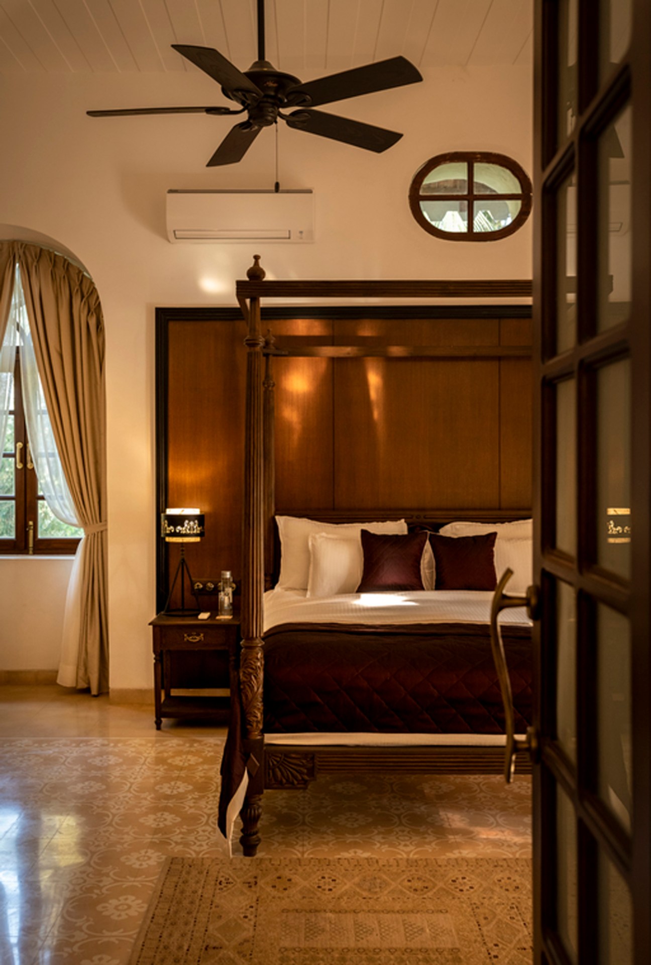 A luxurious bedroom interior featuring a dark wooden four-poster bed with cream and burgundy pillows, a ceiling fan, arched window with golden curtains, and warm ambient lighting typical of upscale hotel design.