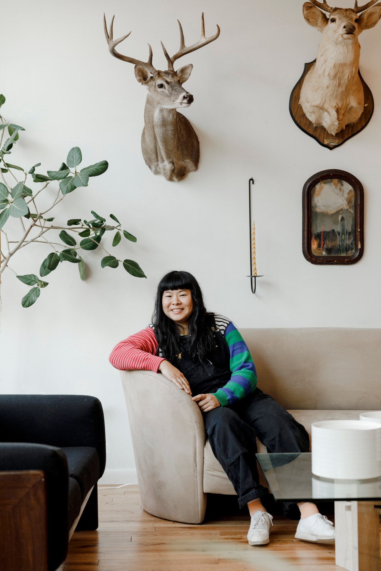 Woman sitting on a beige armchair smiling, surrounded by taxidermy deer heads and vintage décor on white walls.