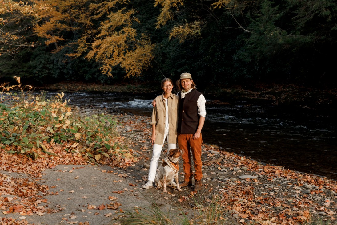 Tom Roberts and Anna Aberg standing by a river in autumn foliage with a dog.